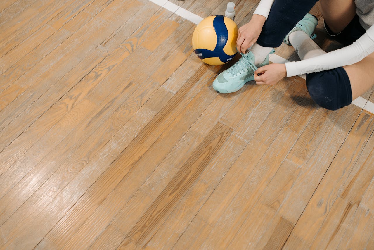 Close-up of an athlete tying shoes on a wooden volleyball court with a ball nearby.